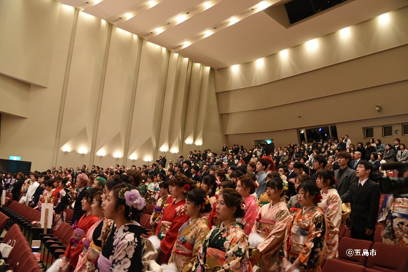 着飾った新成人たちが、文化会館ホールで起立して国歌斉唱している様子。