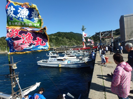 大漁旗を掲げた漁船が港に戻り、もちまきをしている写真です。
